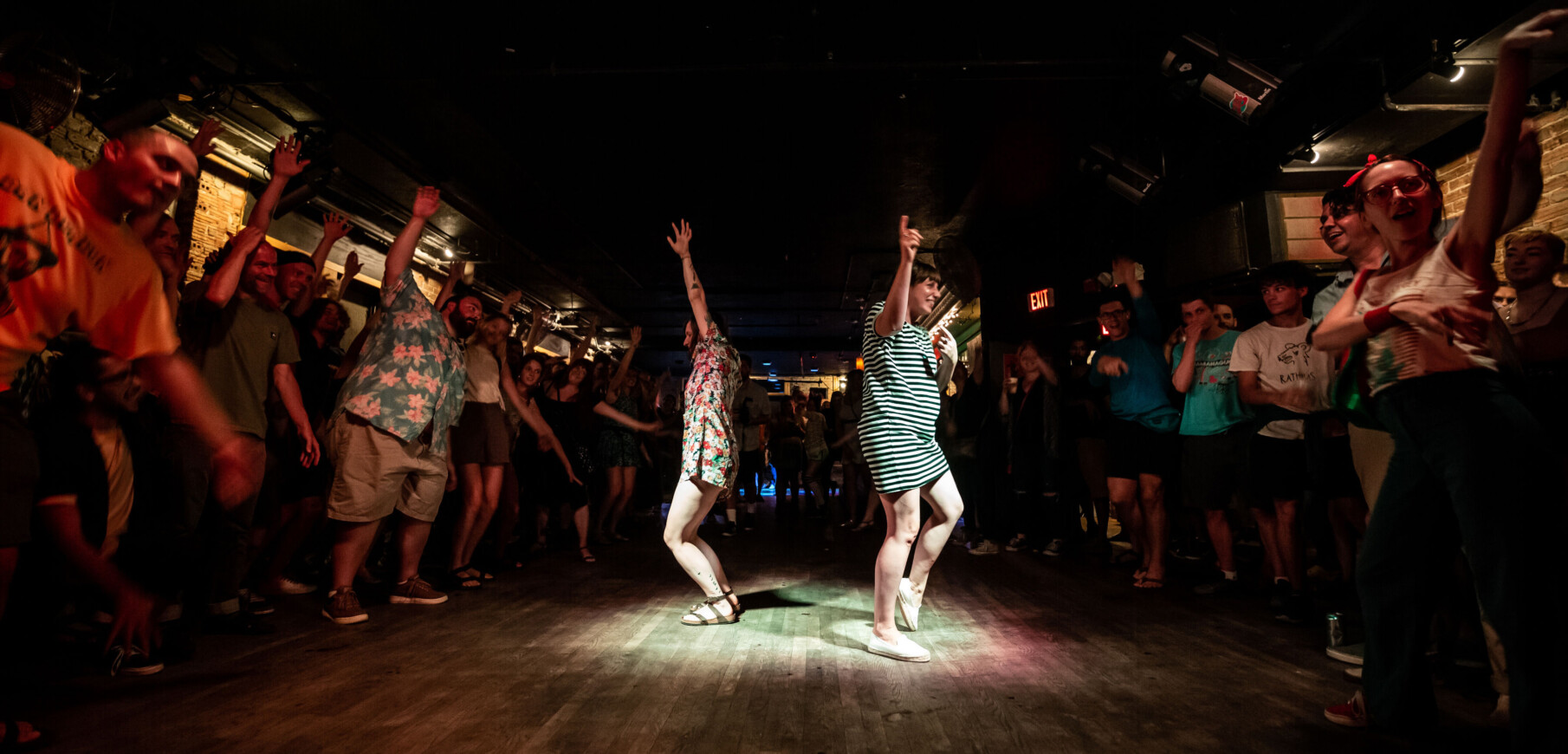 two light skinned women dance under a spotlight as a crowd watches in a darkened room